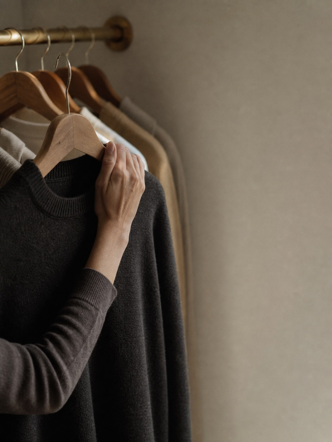 A hand at a closet rail, pulling one hanger forward — a single garment, chosen.