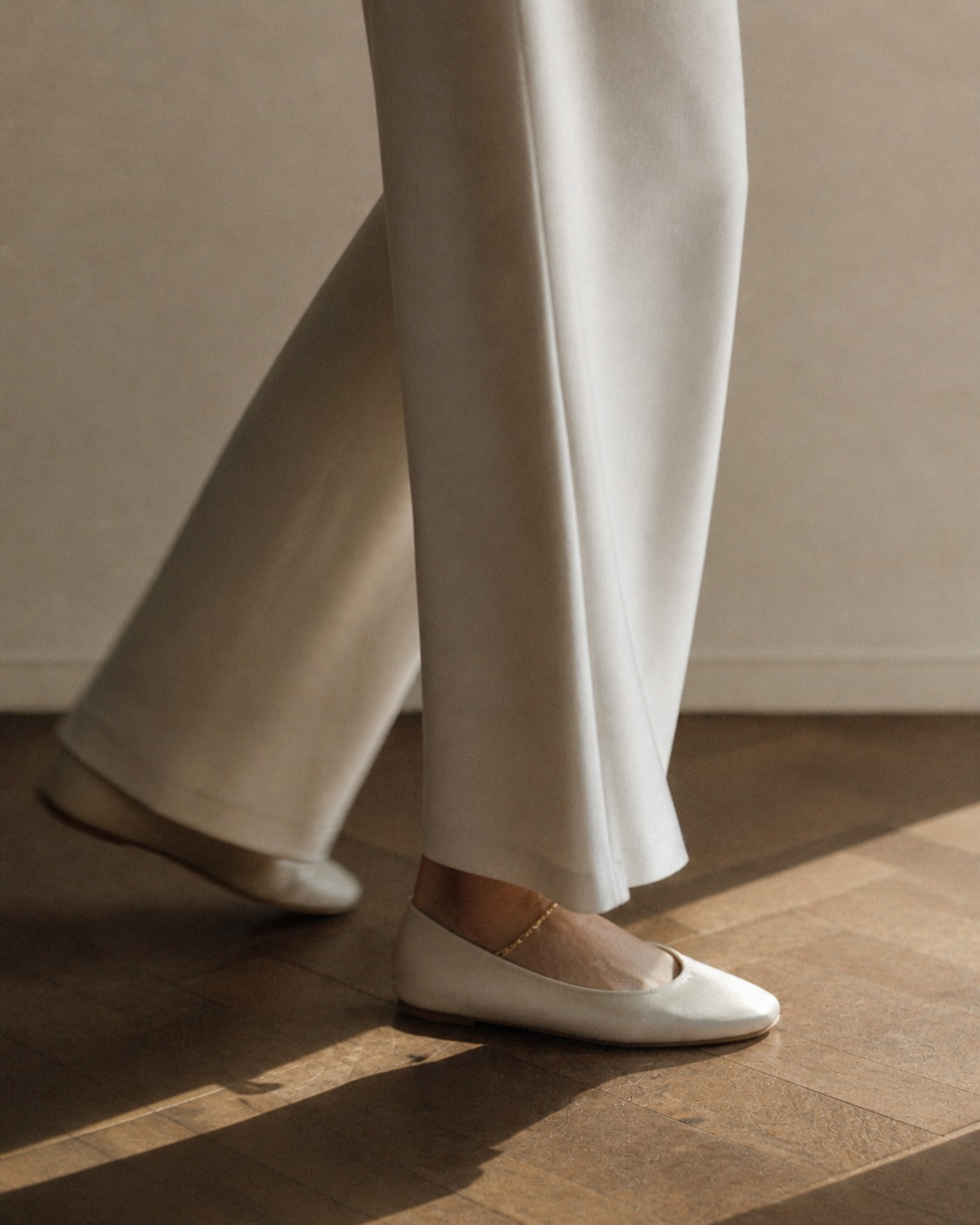 Mid-stride, cream wool trousers and a single gold ankle chain, ivory leather flats.