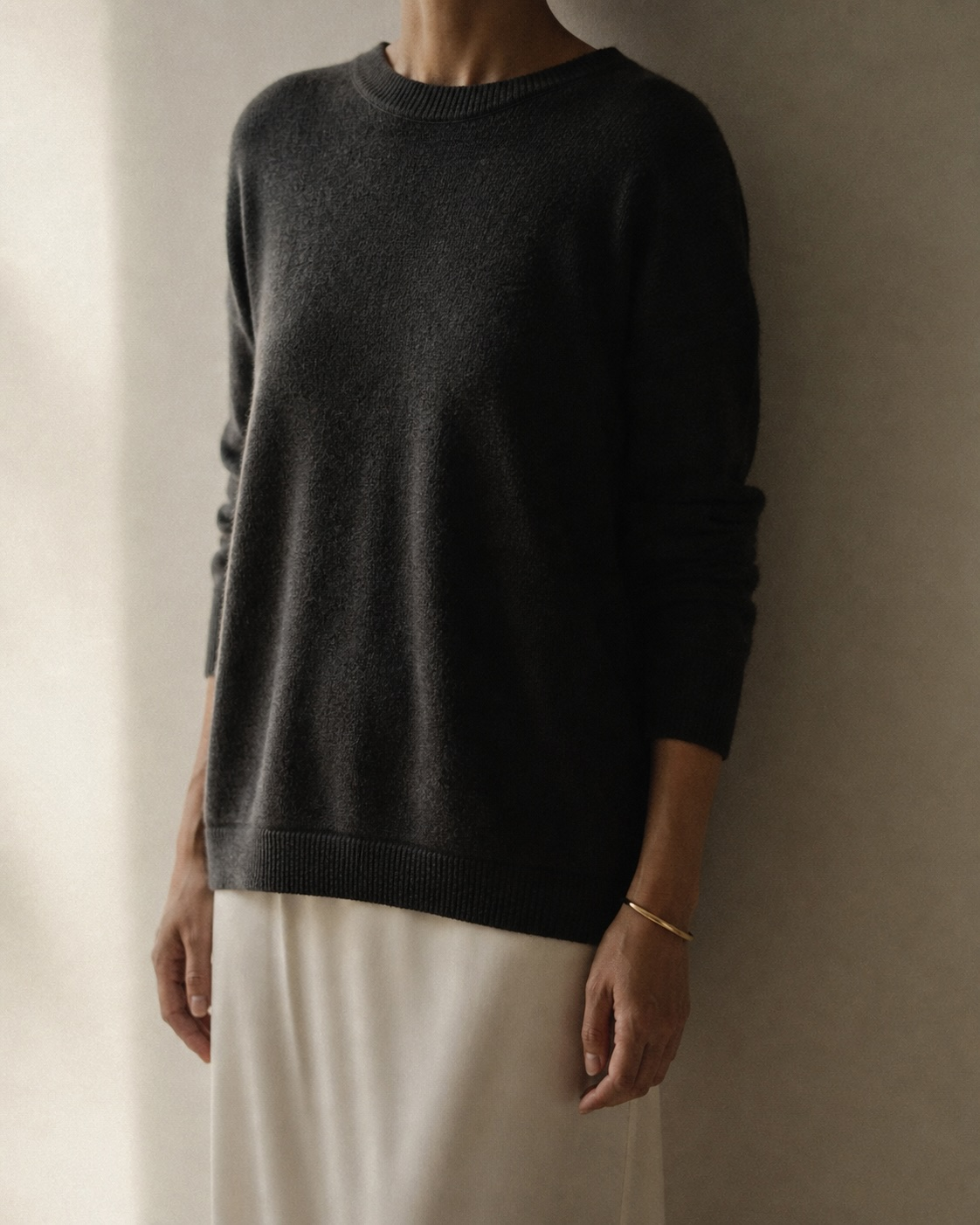 A woman, charcoal cashmere over a silk slip, hands at her sides, window light.